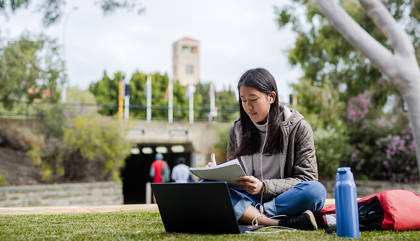 Image of student studying on the grass with laptop with UWA Winthrop Hall in the background